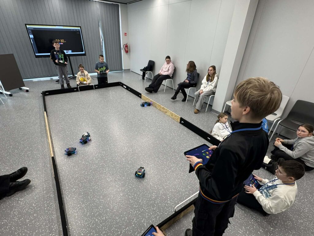 Dzieci siedzące wokół mini boiska i bawiących się tabletami i robotami. 