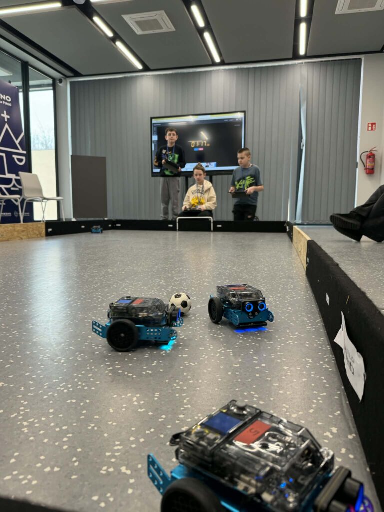 Dzieci siedzące wokół mini boiska i bawiących się tabletami i robotami. 