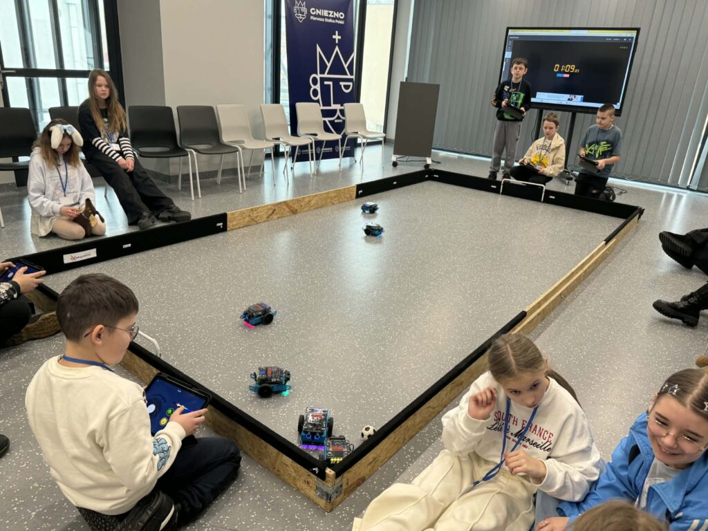 Dzieci siedzące wokół mini boiska i bawiących się tabletami i robotami. 