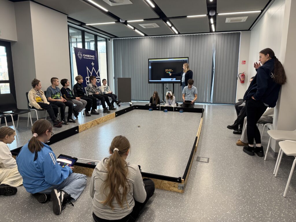 Dzieci siedzące wokół mini boiska i bawiących się tabletami i robotami. 